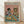 May include: Framed poster print leaning against white wall on light wood floor. Artwork shows anthropomorphic cat in black beret and blue striped shirt sharing brunch with dog in sunglasses and tropical floral shirt. Pink background with teal table displaying croissants, cheese board, baguette, wine glasses, and dog treats. Light wood frame, vintage illustration style.