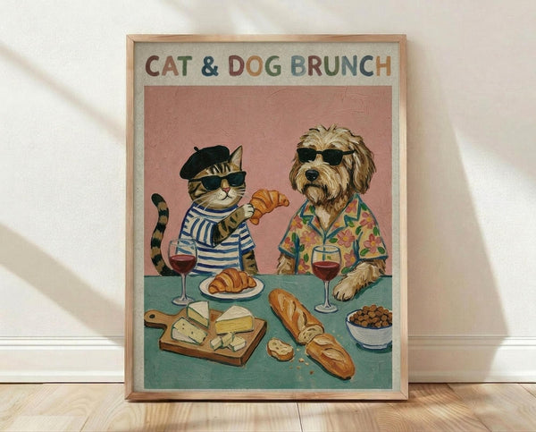 May include: Framed poster print leaning against white wall on light wood floor. Artwork shows anthropomorphic cat in black beret and blue striped shirt sharing brunch with dog in sunglasses and tropical floral shirt. Pink background with teal table displaying croissants, cheese board, baguette, wine glasses, and dog treats. Light wood frame, vintage illustration style.
