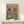 May include: Framed vintage-style poster showing a tabby cat in striped shirt and beret alongside a shaggy dog in Hawaiian shirt, both wearing sunglasses at brunch table with croissants, cheese, wine glasses, and olives against coral pink background. White gift box sits on light wood console table against cream wall.