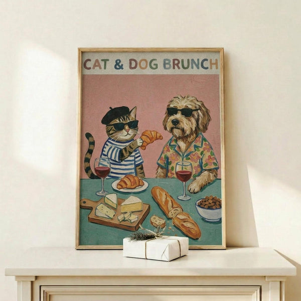 May include: Framed vintage-style poster showing a tabby cat in striped shirt and beret alongside a shaggy dog in Hawaiian shirt, both wearing sunglasses at brunch table with croissants, cheese, wine glasses, and olives against coral pink background. White gift box sits on light wood console table against cream wall.