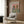May include: Framed poster on wood paneled wall showing anthropomorphic cat in striped shirt and beret alongside shaggy dog in tropical shirt, both wearing sunglasses at brunch table with wine glasses, bread, and cheese. Modern beige armchair with geometric pillow, gold floor lamp, and dark round side table with open book in contemporary room setting.