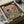 May include: Framed vintage-style poster illustration showing a tabby cat in striped shirt and beret alongside a Yorkshire terrier in tropical Hawaiian shirt, both wearing sunglasses at brunch table with croissants, wine glasses, cheese board and baguette. Artwork has coral pink background with teal table, packed in cardboard box with protective wrapping materials.