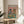 May include: Framed poster print on wooden side table next to beige upholstered chair and plant. Artwork shows anthropomorphic cat in striped shirt and beret with dog in floral shirt, both wearing sunglasses at brunch table with croissants, cheese, bread, and wine glasses. Pink and teal color scheme with natural wood frame.