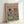 Framed poster featuring a stylized cat in beret and striped shirt alongside a shaggy dog in tropical shirt, both wearing sunglasses at brunch table with croissants, wine glasses, cheese board and bread. Pink background with teal table surface. Light wood frame leaning against white wall.