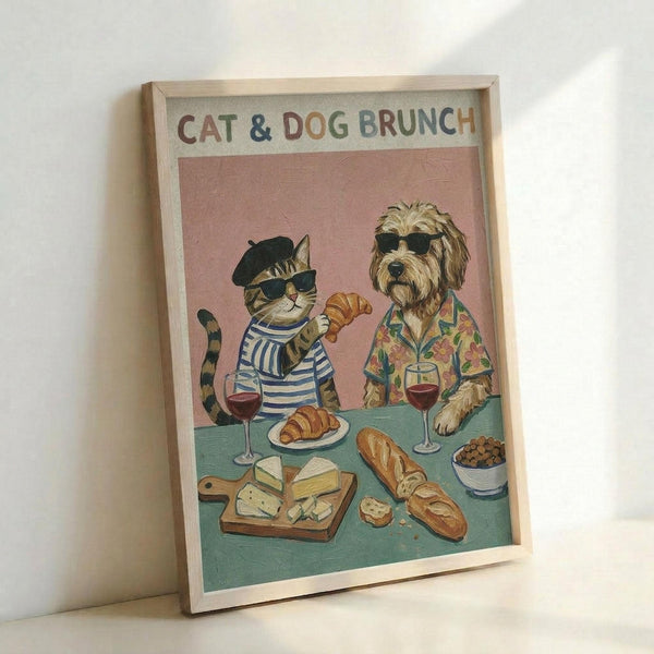 Framed poster featuring a stylized cat in beret and striped shirt alongside a shaggy dog in tropical shirt, both wearing sunglasses at brunch table with croissants, wine glasses, cheese board and bread. Pink background with teal table surface. Light wood frame leaning against white wall.