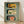 May include: Three vintage-style Portuguese sardine can labels displayed in a wooden frame against a sage green background. The framed artwork sits on a light wooden floor against a white wall. Labels show "Tricana" with a woman's portrait, "Prata Do Mar" with a green sailboat, and "Minor" with a red cat mascot, all featuring retro typography and nautical themes.