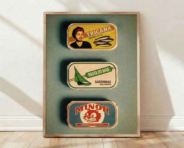 May include: Three vintage-style Portuguese sardine can labels displayed in a wooden frame against a sage green background. The framed artwork sits on a light wooden floor against a white wall. Labels show "Tricana" with a woman's portrait, "Prata Do Mar" with a green sailboat, and "Minor" with a red cat mascot, all featuring retro typography and nautical themes.