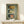 May include: Framed vintage-style poster print featuring three retro sardine tin can designs - yellow Tricana with woman portrait, white Prata Do Mar with green sailboat, and teal Minor with red cat mascot. Displayed on light wood console with small wrapped gift, against cream wall with natural lighting.