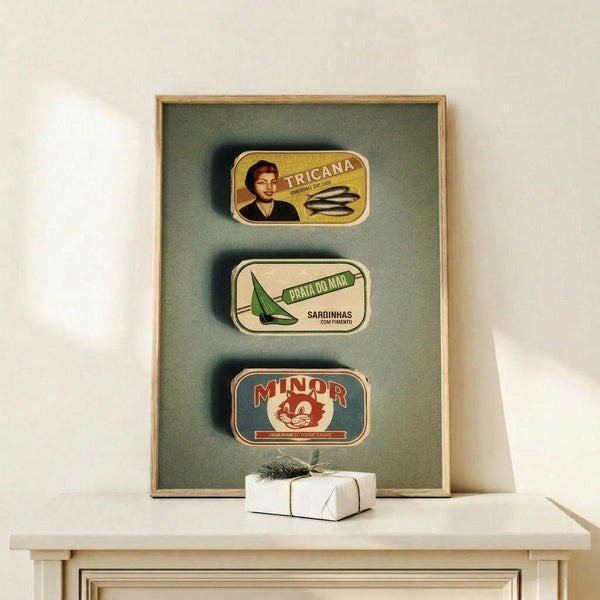 May include: Framed vintage-style poster print featuring three retro sardine tin can designs - yellow Tricana with woman portrait, white Prata Do Mar with green sailboat, and teal Minor with red cat mascot. Displayed on light wood console with small wrapped gift, against cream wall with natural lighting.