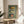 May include: Three vintage-style Portuguese sardine can labels displayed in a light wood frame against sage green background. The artwork shows colorful retro packaging designs including Tricana, Prata do Mar, and Minor brands. Frame sits on round wooden side table next to beige upholstered chair with white pillow, palm plant visible on left, soft natural lighting creates shadows on white wall.