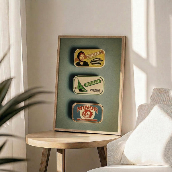 May include: Three vintage-style Portuguese sardine can labels displayed in a light wood frame against sage green background. The artwork shows colorful retro packaging designs including Tricana, Prata do Mar, and Minor brands. Frame sits on round wooden side table next to beige upholstered chair with white pillow, palm plant visible on left, soft natural lighting creates shadows on white wall.