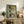May include: Framed vintage-style poster displaying three Portuguese sardine can labels in yellow, white, and red colors mounted on sage green background. Wooden frame leans against white wall on rustic wood credenza beside fern plant, books, and ceramic vase in neutral-toned living room setting.