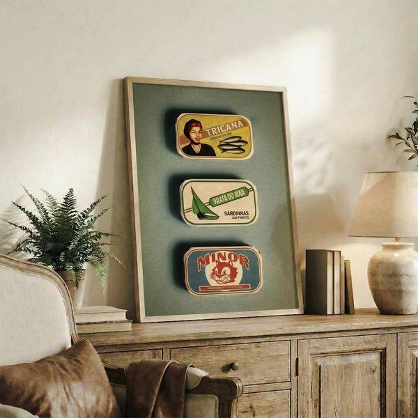 May include: Framed vintage-style poster displaying three Portuguese sardine can labels in yellow, white, and red colors mounted on sage green background. Wooden frame leans against white wall on rustic wood credenza beside fern plant, books, and ceramic vase in neutral-toned living room setting.