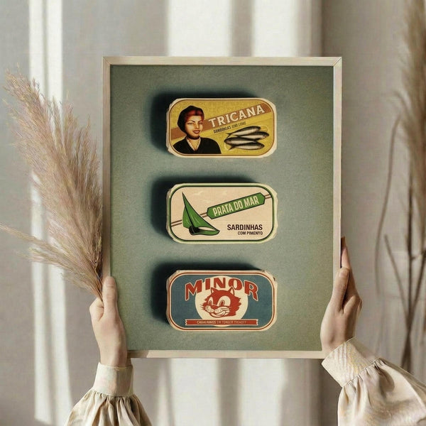 May include: Hands holding framed vintage sardine can label artwork against sage green background. Three colorful retro Portuguese canned fish labels mounted in light wood frame - yellow Tricana with woman portrait, white Praia do Mar with green leaf design, and teal Minor with cat illustration. Dried pampas grass visible on left side.