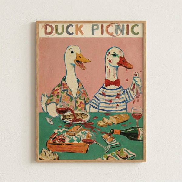 Whimsical illustration of two anthropomorphic ducks having a picnic, framed in light wood against a white wall. The left duck wears a floral shirt while the right duck sports a striped top and red bow tie, with red hair and makeup giving a feminine appearance. Both sit at a green picnic table covered with food including fish, bread, wine glasses, and a bottle. The artwork features a pink background with colorful, playful styling reminiscent of vintage children's book illustrations.