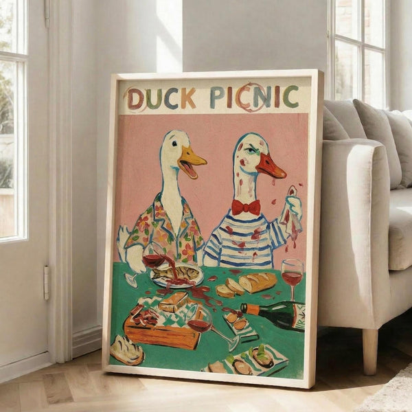 May include: Framed poster print leaning against white chair in bright living room with windows. Artwork shows two anthropomorphic ducks in human clothing having a picnic at green table with food, wine, and bread. Pink background with colorful text reading "DUCK PICNIC" at top. Light wood frame on hardwood floor with natural lighting.