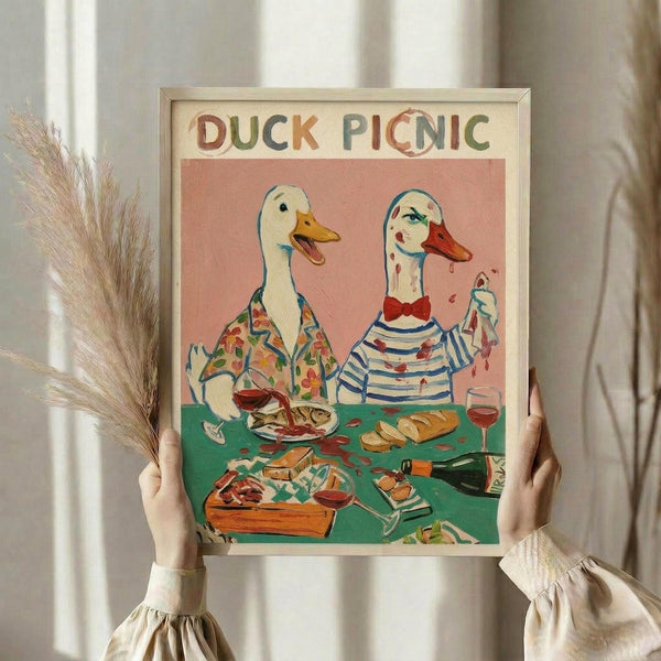 May include: Person in cream ruffled sleeves holding framed poster print and dried wheat stems against white curtained window. Artwork shows two anthropomorphic ducks having a picnic - one in floral shirt, one in striped shirt with red bow tie, sitting at green table with wine, bread, and food. Pink background with colorful, whimsical illustration style in light wooden frame.