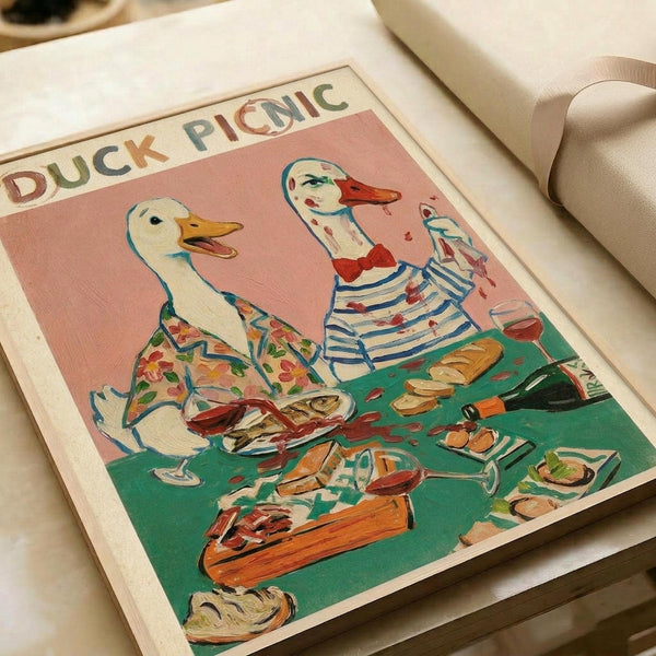 Two anthropomorphic ducks in vintage clothing sit at a green picnic table with wine glasses, bread, and food. The left duck wears a floral shirt, the right duck sports a striped top and red bow tie. Set against a coral pink background, this whimsical illustration has a retro poster aesthetic with "DUCK PICNIC" text at the top. The artwork appears as a print displayed on white paper with cream ribbon visible in the corner.