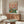 May include: Whimsical poster artwork featuring two anthropomorphic ducks in clothing sitting at a green picnic table with food and wine, displayed above a mid-century modern walnut credenza in a minimalist living room with white walls, cream chair, glass coffee table, and warm natural lighting