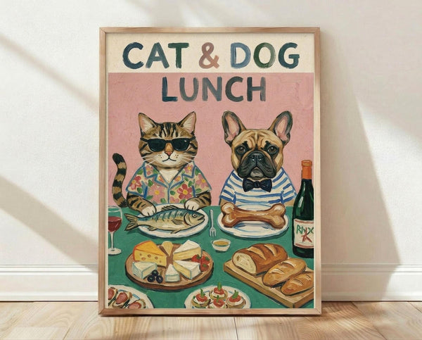 May include: Framed poster print leaning against white wall on light wood flooring. Artwork shows anthropomorphized cat in sunglasses and floral shirt sitting with French bulldog in striped shirt at lunch table. Cat has fish on plate, dog has bone. Table displays cheese platter, bread, wine bottle, and appetizers on green tablecloth. Pink background with cream header reading "CAT & DOG LUNCH" in green letters. Natural wood frame.
