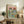 May include: Framed poster displayed on rustic wooden sideboard in cozy living room setting. Artwork features two cats wearing sunglasses and hats at a picnic with fish, wine, and food items against pink and green background. Colorful lettering at top reads "PURR-FECT PICNIC". Natural wood frame, surrounded by fern plant, books, ceramic vase, and beige furniture.