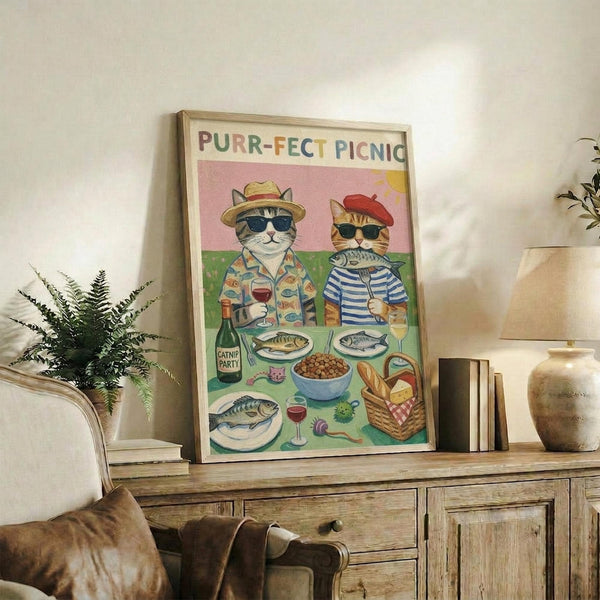 May include: Framed poster displayed on rustic wooden sideboard in cozy living room setting. Artwork features two cats wearing sunglasses and hats at a picnic with fish, wine, and food items against pink and green background. Colorful lettering at top reads "PURR-FECT PICNIC". Natural wood frame, surrounded by fern plant, books, ceramic vase, and beige furniture.