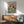 May include: Framed poster displaying illustrated cat wearing sunglasses and Hawaiian shirt sitting beside French bulldog in striped shirt at dining table with fish, bone, cheese, bread and wine. Shown mounted above wood credenza with white lamp in modern living room setting with mint green armchair.