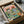 May include: Framed vintage-style illustration of two cats wearing sunglasses and hats at a picnic, surrounded by fish dishes, wine, and food. The colorful poster features "PURR-FECT PICNIC" text at top with pink sky background. Black frame sits in cardboard shipping box with white tissue paper wrapping, alongside rolled tube packaging materials.