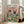 May include: Framed poster artwork leaning against white wall next to beige sofa in bright room with large windows. Print shows anthropomorphic tabby cat wearing sunglasses and floral shirt sitting at green table with French bulldog in striped shirt and bow tie. Table spread includes fish, bone, cheese platter, bread, wine bottle and various foods. Pink and cream background with teal lettering at top. Light wood frame, approximately 2-3 feet tall.
