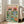 May include: Framed poster print leaning against white wall next to beige sofa in bright living room. Artwork shows two cats wearing sunglasses and hats at colorful picnic table with fish dishes, wine, bread basket, and cat toys. Features pink sky background, green grass, and "PURR-FECT PICNIC" text in rainbow letters across top. Light wooden frame, approximately 2-3 feet tall.