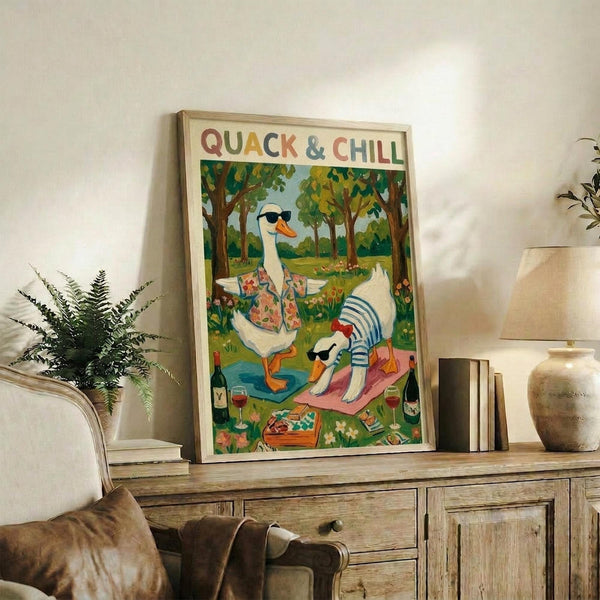 May include: Framed poster showing two white ducks with sunglasses relaxing outdoors - one wearing a floral shirt standing by water, another in striped shirt lying on a pink blanket. Colorful cartoon style with green trees, wine bottles and picnic items. Natural wood frame displayed on rustic wooden sideboard with potted fern, ceramic vases, books, and brown leather chair visible in cozy living room setting with white walls.