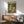 May include: Framed poster showing cartoon duck in sunglasses and Hawaiian shirt with cat in striped shirt having a picnic in green park setting. Wood sideboard with white ceramic lamp below, teal upholstered mid-century armchair beside. Bright sunlight streaming across white wall. Warm wood tones throughout furniture pieces.