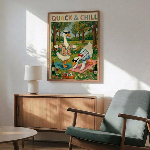 May include: Framed poster showing cartoon duck in sunglasses and Hawaiian shirt with cat in striped shirt having a picnic in green park setting. Wood sideboard with white ceramic lamp below, teal upholstered mid-century armchair beside. Bright sunlight streaming across white wall. Warm wood tones throughout furniture pieces.