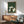May include: Framed poster print hanging on white wall above wood sideboard with table lamp. Green textured background artwork shows five cats sitting around dark wooden table playing cards. Cats include white, black, and mixed colors in Japanese woodblock print style. Mid-century modern room with teal upholstered chair, round coffee table. Natural sunlight creates shadows on wall.