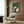 May include: Framed poster with green background showing white and black cats gathered around dark wooden table playing cards, displayed on wood paneled wall above beige upholstered armchair with geometric patterned pillow, brass floor lamp and side table with open book nearby