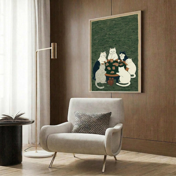 May include: Framed poster with green background showing white and black cats gathered around dark wooden table playing cards, displayed on wood paneled wall above beige upholstered armchair with geometric patterned pillow, brass floor lamp and side table with open book nearby