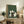 May include: Framed artwork displaying four cats sitting around a dark wooden table playing cards, rendered in Japanese woodblock print style with green textured background. The cats include white, black, and gray varieties. The wooden-framed piece sits on a rustic sideboard alongside potted plants, books, and ceramic vessels in a neutral-toned living room setting.