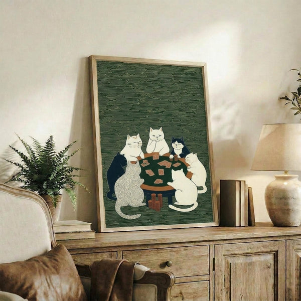 May include: Framed artwork displaying four cats sitting around a dark wooden table playing cards, rendered in Japanese woodblock print style with green textured background. The cats include white, black, and gray varieties. The wooden-framed piece sits on a rustic sideboard alongside potted plants, books, and ceramic vessels in a neutral-toned living room setting.