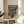 May include: A framed vintage-style sci-fi poster displayed on a wooden round side table next to a beige upholstered chair. The artwork features a person in an orange spacesuit standing before a large mech robot, with Japanese text at the top and teal-green atmospheric coloring throughout. Natural sunlight streams across the minimalist room setting with white walls and a small plant visible in the corner.