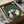 May include: Framed artwork featuring six cats in various colors (white, black, striped) sitting around a dark wooden table playing cards, rendered in Japanese woodblock print style with green textured background. The piece is being unpacked from a cardboard shipping box with protective wrapping, showing the wooden frame and matting details.