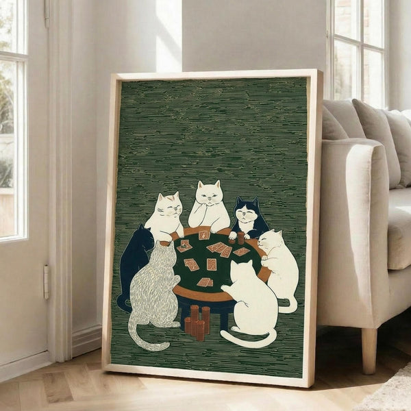 May include: Framed artwork showing five cats gathered around a green poker table playing cards, rendered in Japanese woodblock print style with green textured background. The cats are various colors - white, black, and striped - sitting upright like humans. Brown poker chips are stacked nearby. The piece is displayed in a light wood frame, leaning against a beige armchair in a bright, windowed living room with white walls and wooden floors.