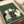 May include: Green woodblock-style print showing five cats playing poker around a wooden table with red playing cards and poker chips scattered on top. The cats are rendered in white, black, and striped patterns against a textured green background with fine horizontal lines. The artwork is displayed flat on white paper with a light wooden frame border, positioned next to a white cylindrical object on what appears to be a light wooden surface.