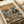 May include: Vintage-style poster print displayed on white bedding showing two koalas in sunglasses dining at a wooden table by the ocean at sunset. One koala wears a floral Hawaiian shirt, the other a black tuxedo. The table features a whole cooked fish, wine glasses, cheese, and eucalyptus leaves. Text reads "KOALA FINE DINING" at top. Warm orange and yellow sunset sky with ocean waves in background. Print appears flat against white fabric surface.