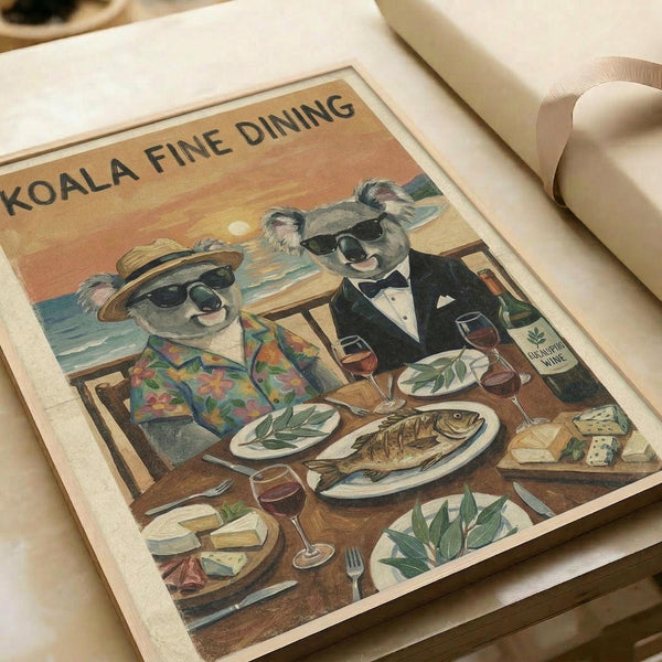 May include: Vintage-style poster print displayed on white bedding showing two koalas in sunglasses dining at a wooden table by the ocean at sunset. One koala wears a floral Hawaiian shirt, the other a black tuxedo. The table features a whole cooked fish, wine glasses, cheese, and eucalyptus leaves. Text reads "KOALA FINE DINING" at top. Warm orange and yellow sunset sky with ocean waves in background. Print appears flat against white fabric surface.