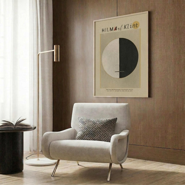 May include: Framed geometric art print featuring a circle split into cream and dark green halves with "HILMA af Klint" text, displayed on wood paneled wall above a beige upholstered armchair with patterned throw pillow, brass floor lamp and dark round side table with open book nearby