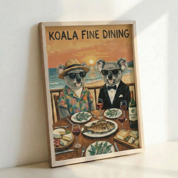 A framed vintage-style poster showing two koalas in sunglasses dining at a wooden table overlooking a beach sunset. One wears a tropical shirt and hat, the other a black tuxedo with bow tie. The table displays wine glasses, eucalyptus leaves on plates, fish, cheese, and a wine bottle. The artwork features warm orange and yellow sunset colors against blue ocean waves. The poster is displayed in a light wood frame leaning against a white wall.