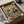 May include: Vintage-style poster print in black frame being unboxed from cardboard packaging with protective tissue paper. Artwork shows two koalas wearing sunglasses at seaside dinner table - one in floral Hawaiian shirt and hat, other in black tuxedo. Table set with grilled fish, wine glasses, cheese and crackers against orange sunset ocean backdrop. Warm coral and teal color palette with retro illustration style.