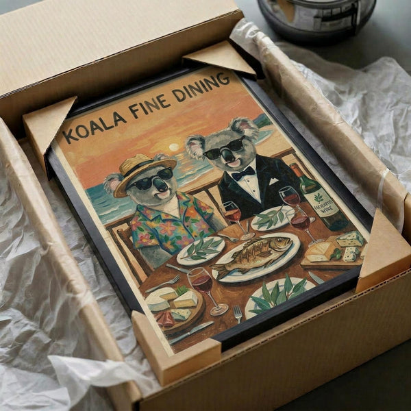 May include: Vintage-style poster print in black frame being unboxed from cardboard packaging with protective tissue paper. Artwork shows two koalas wearing sunglasses at seaside dinner table - one in floral Hawaiian shirt and hat, other in black tuxedo. Table set with grilled fish, wine glasses, cheese and crackers against orange sunset ocean backdrop. Warm coral and teal color palette with retro illustration style.