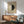 May include: Framed Hilma af Klint exhibition poster featuring a large circle split into white and black halves on beige background, displayed above a wooden sideboard with table lamp in modern living room setting with mint green armchair and natural sunlight casting shadows on white wall