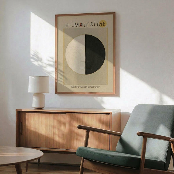 May include: Framed Hilma af Klint exhibition poster featuring a large circle split into white and black halves on beige background, displayed above a wooden sideboard with table lamp in modern living room setting with mint green armchair and natural sunlight casting shadows on white wall